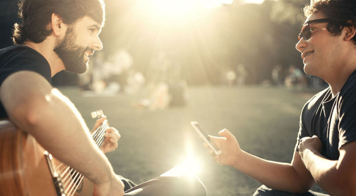 Männer sitzen im Park mit Gitarre und Smartphone.