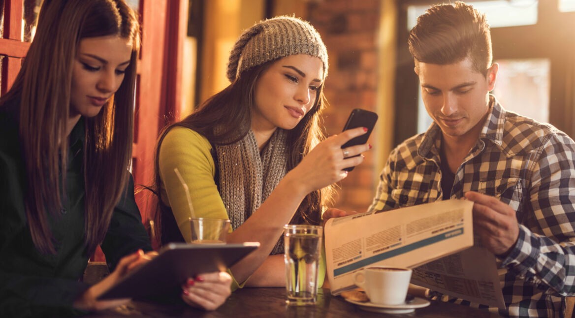Freunde sitzen an einem Tisch, spielen mit dem Smartphone und Tablet, lesen eine Zeitung.