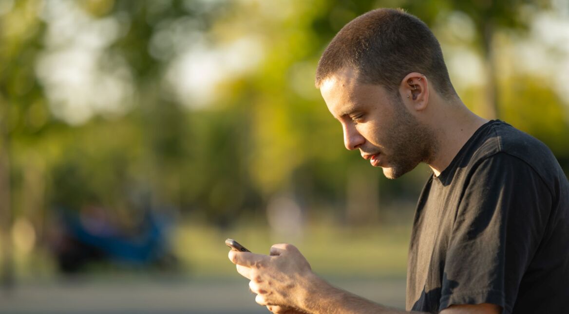 Junger Mann sitzt im Park und schaut auf sein Android-Handy.