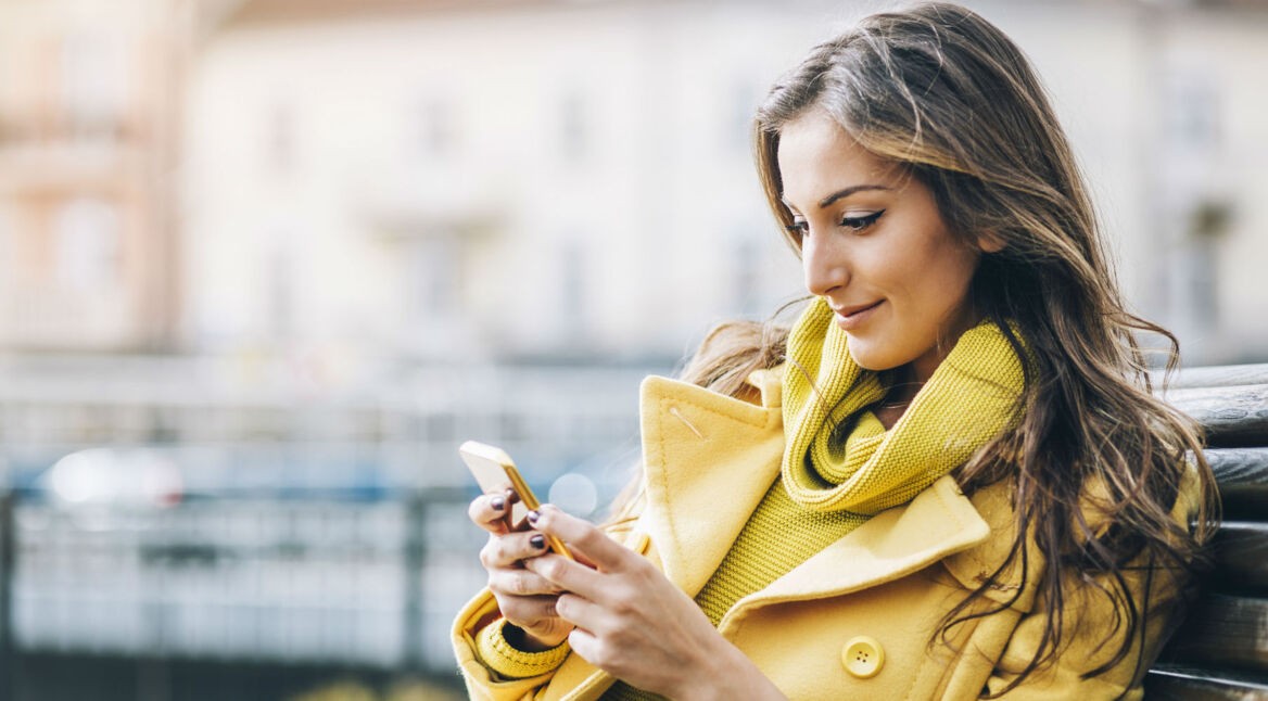 Frau in gelbem Mantel sitzt auf einer Bank mit ihrem Smartphone in der Hand.