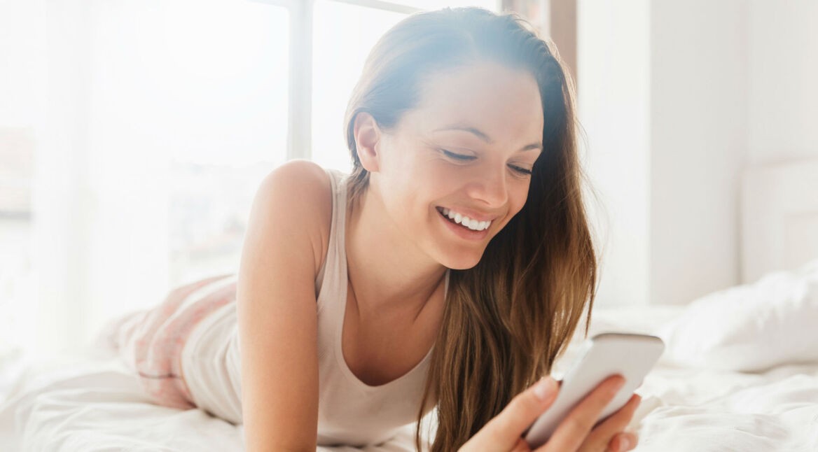 Frau liegt im Bett und schaut auf ihr Smartphone.