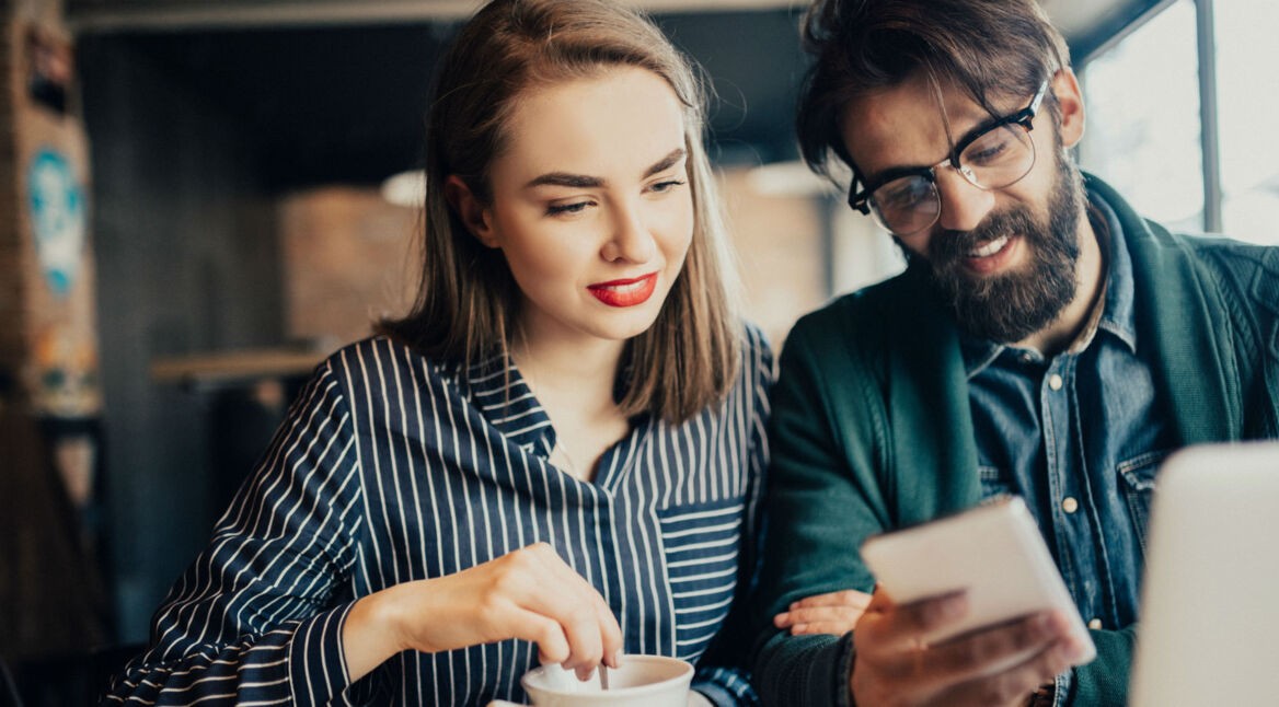 Frau und Mann schauen auf ein Smartphone, während sie in der Kaffeetasse rührt.