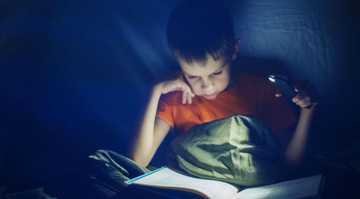 Junge liest nachts im Zelt ein Buch mit Smartphone-Taschenlampe.