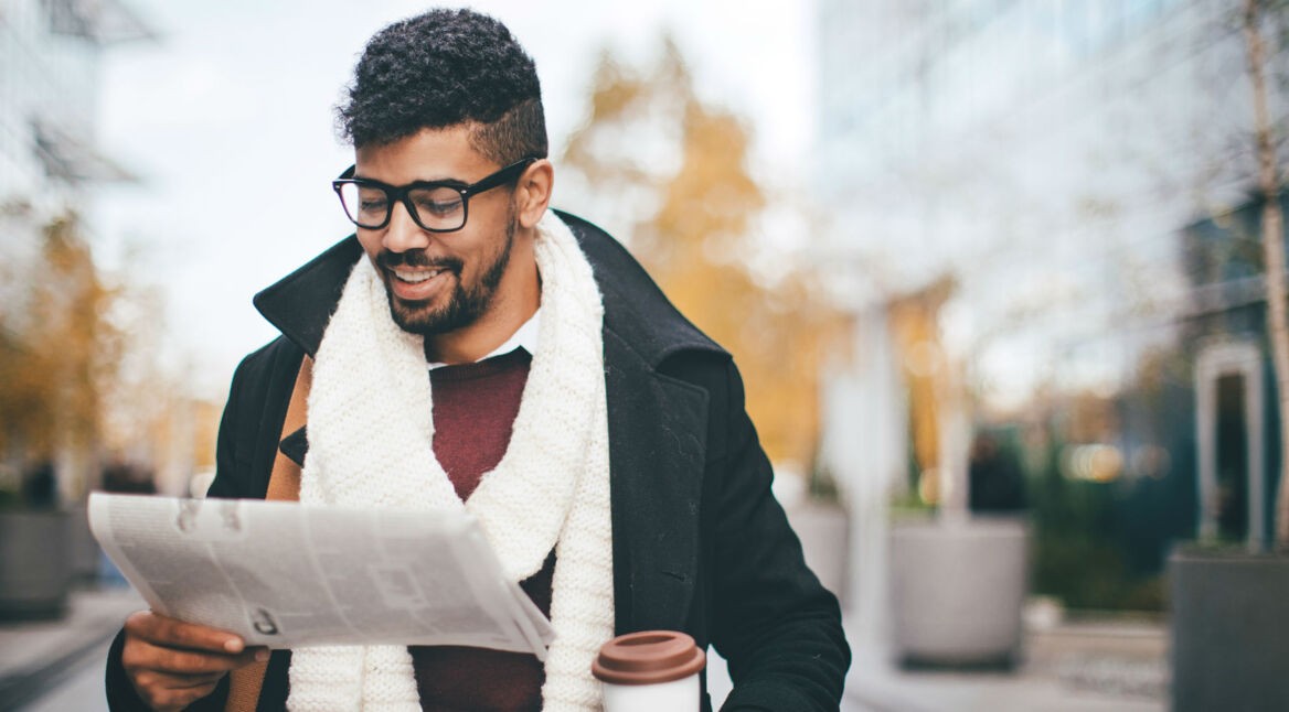Junger Mann liest Zeitung und hält Kaffeebecher in der Hand.