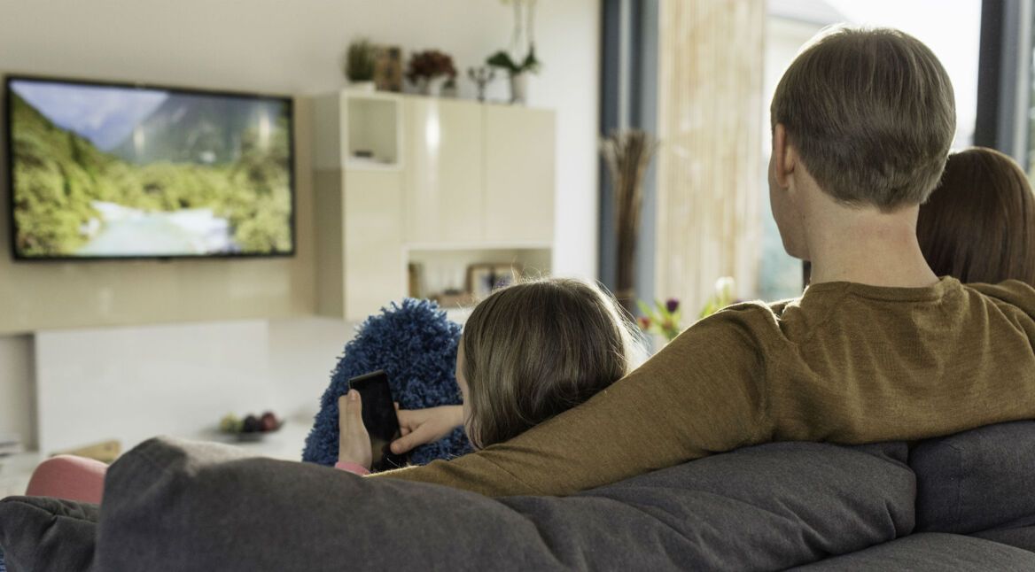Familie sitzt vor dem Fernseher auf dem heimischen Sofa.