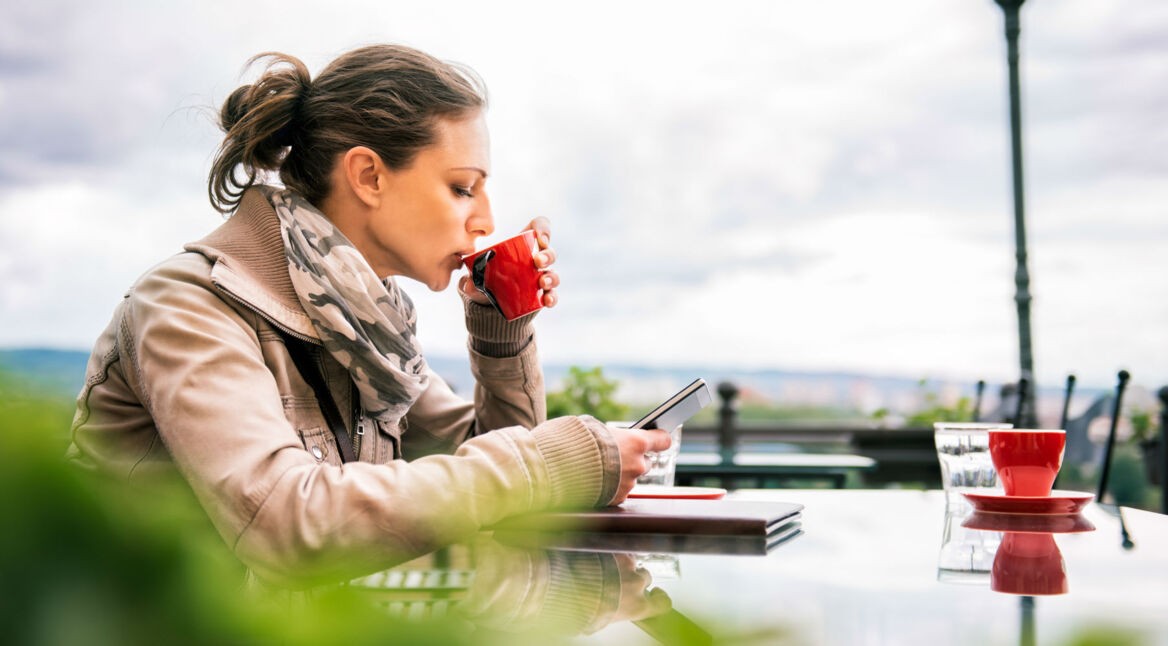 Frau trinkt Kaffee aus einer roten Tasse und schaut auf ihr Smartphone.