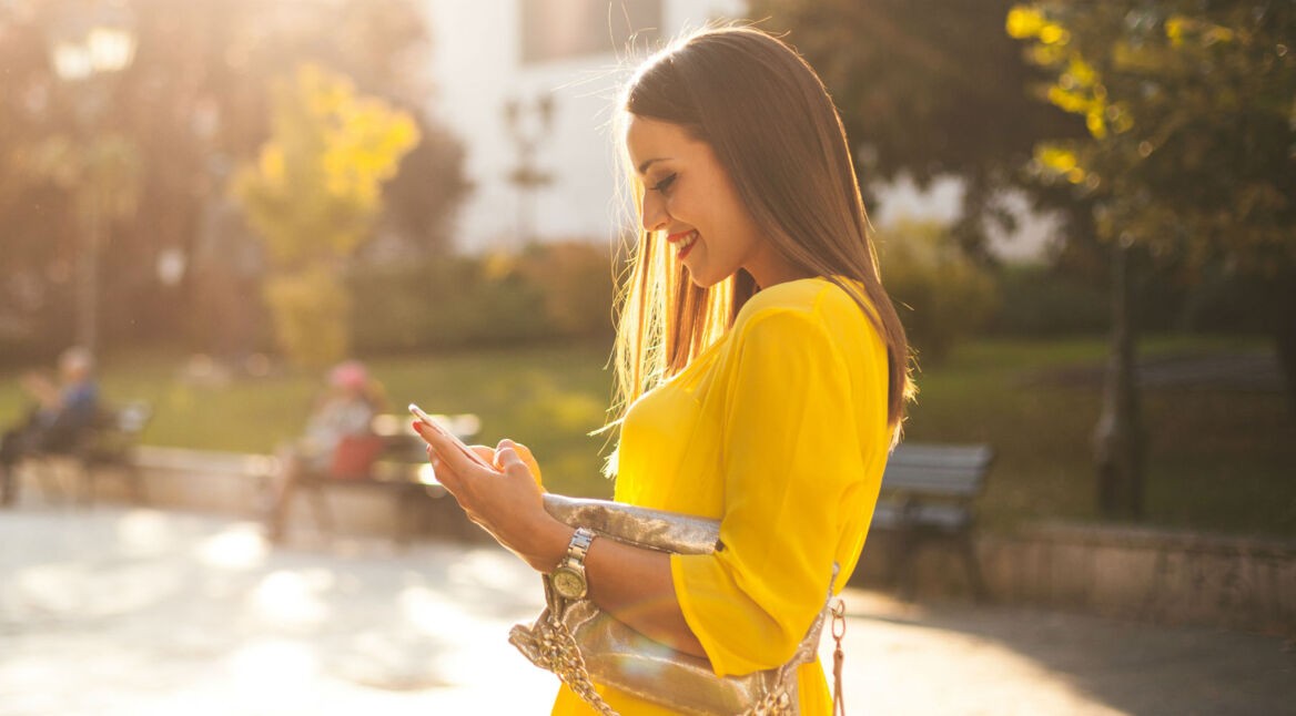 Junge Frau in gelbem Kleid hält Smartphone in der Hand und lächelt.