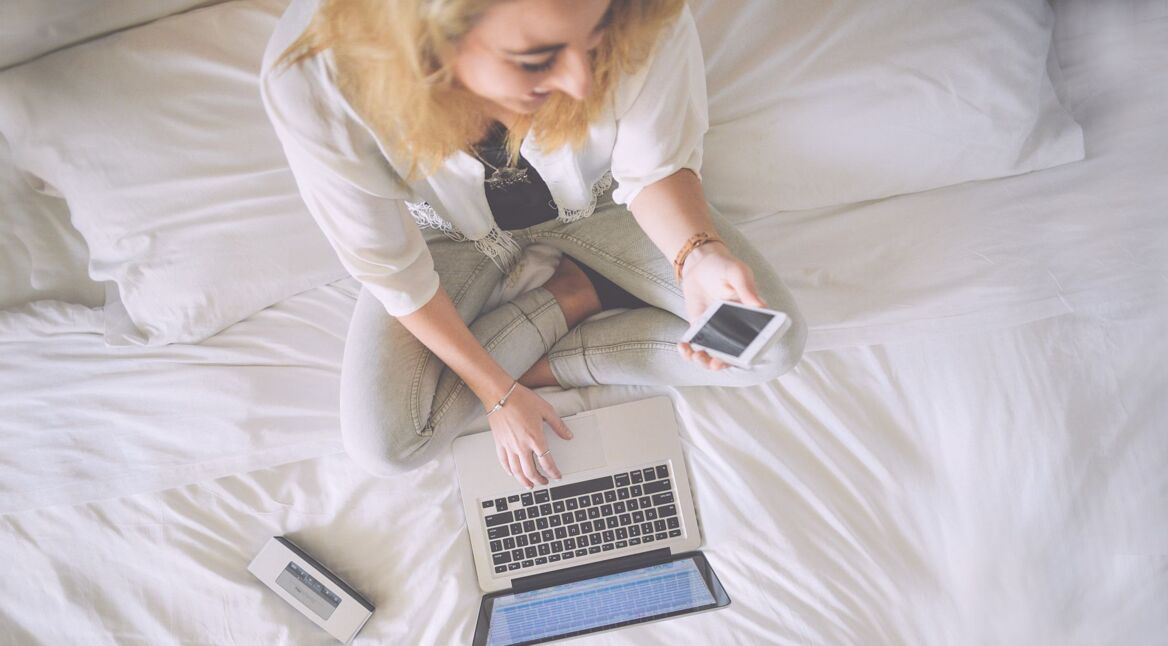 Lachende Frau auf Bett mit iPhone und MacBook.