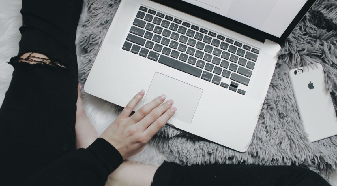 Junge Frau tippt auf Macbook, neben ihr liegt ein iPhone auf dem Bett.