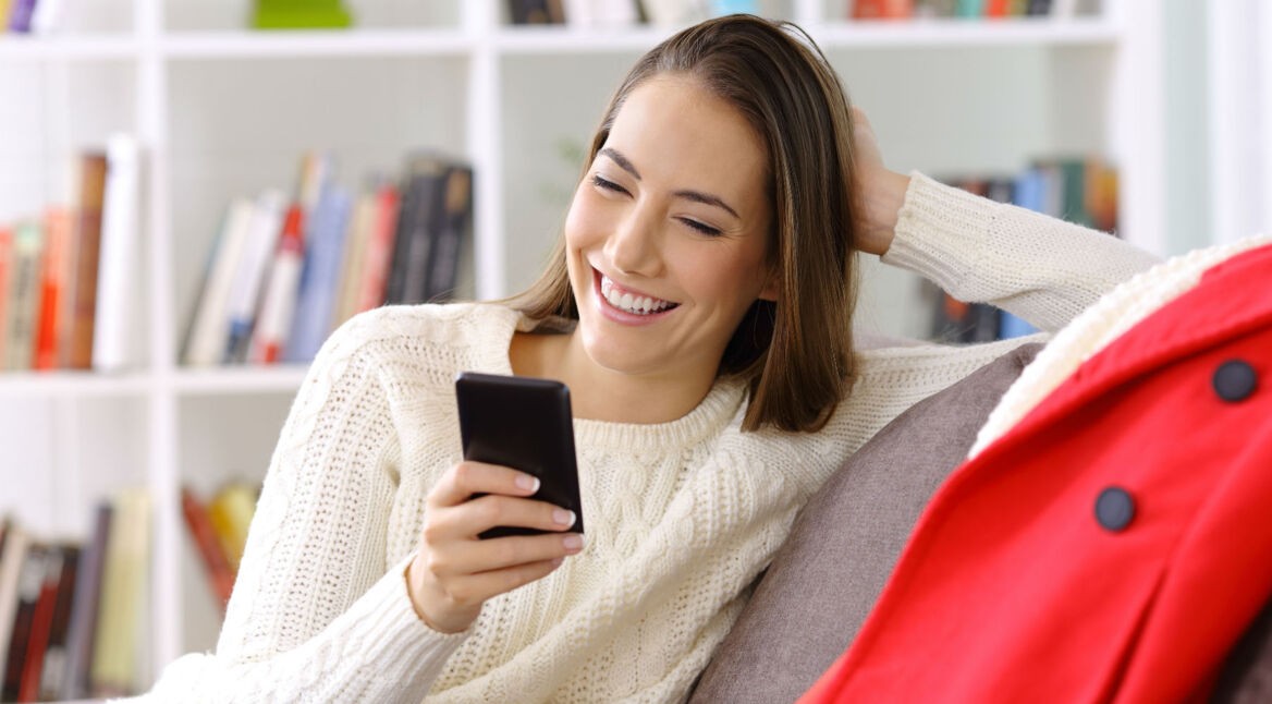 Junge Frau sitzt mit ihrem Smartphone in der Hand auf dem Sofa.