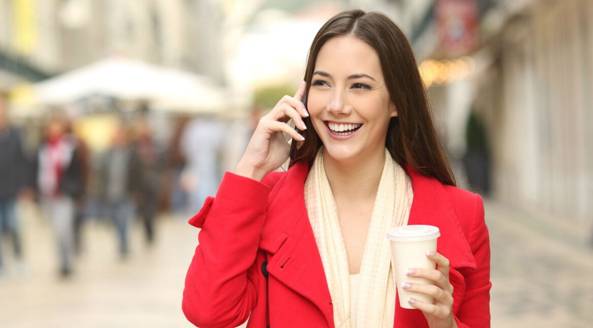 Junge Frau in rotem Mantel telefoniert und hält Kaffeebecher in der Hand.