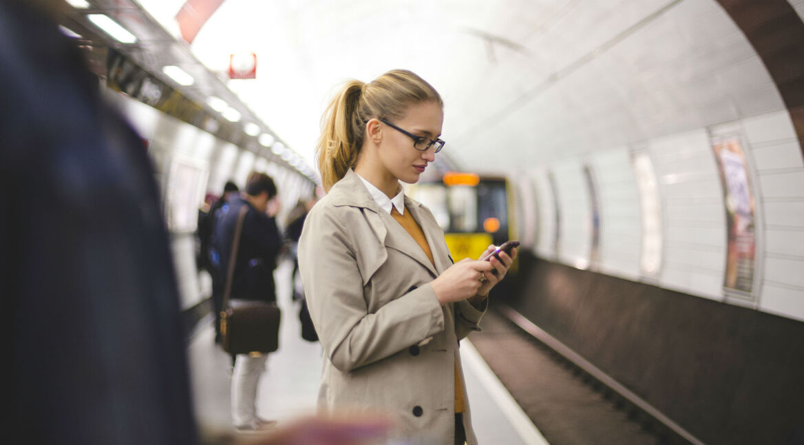 Frau steht am Bahnsteig und surft am Smartphone.
