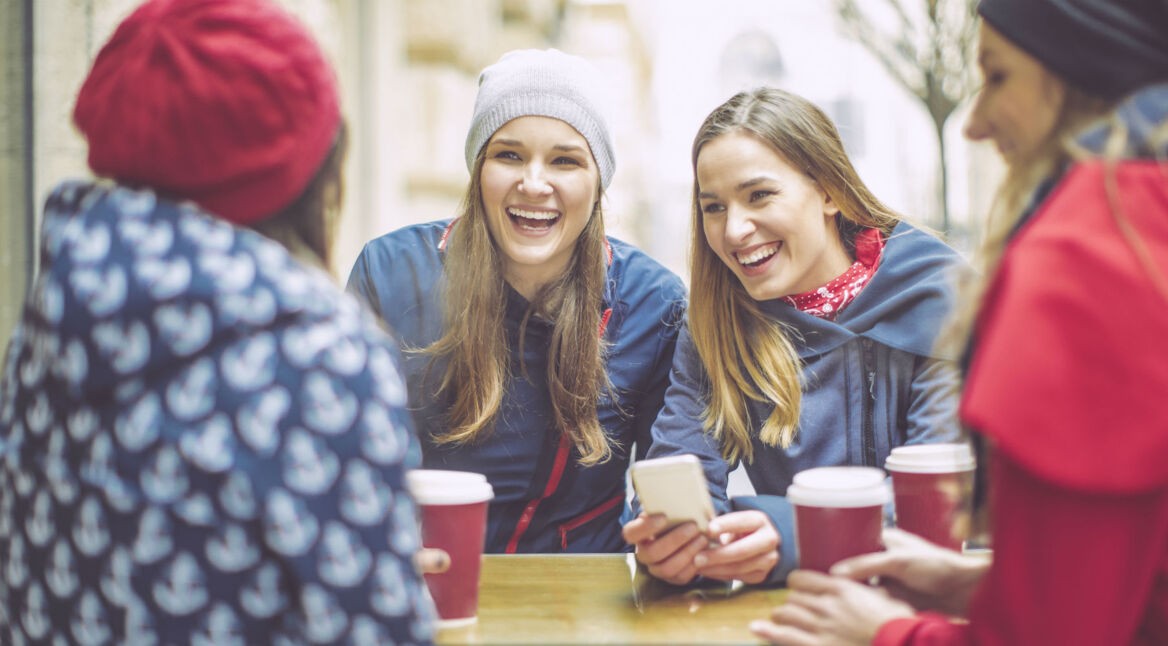 Vier Freundinnen unterhalten sich am Tisch, eine hält ihr Smartphone in der Hand.