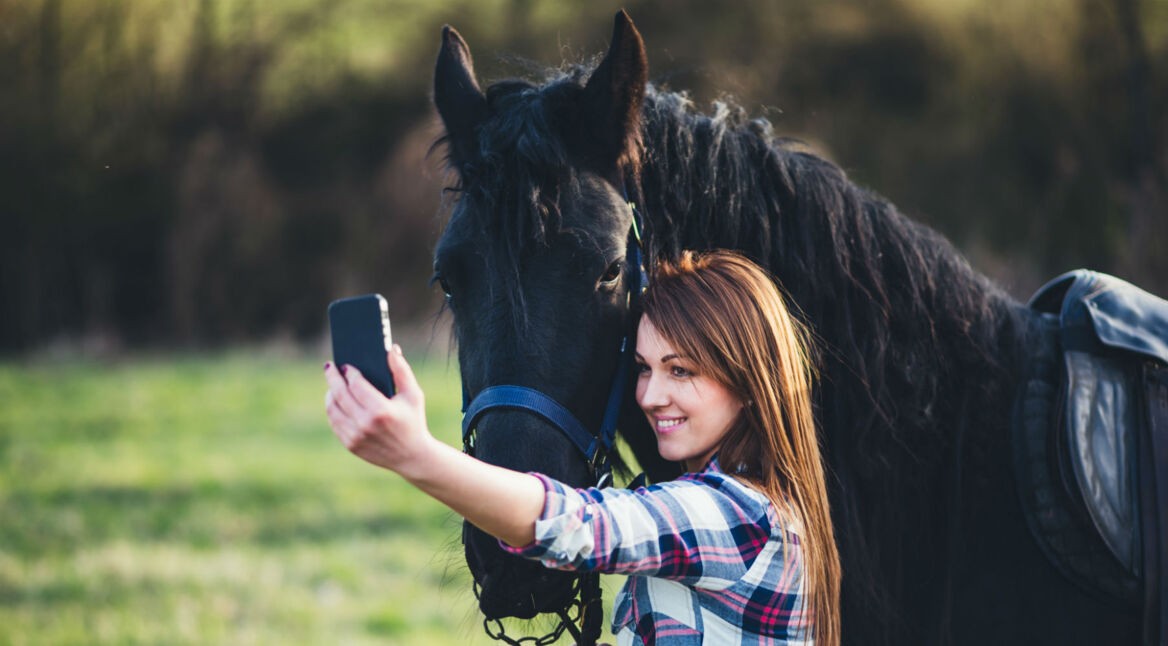 Mädchen posiert neben ihrem Pferd mit dem Smartphone.