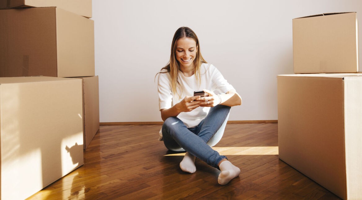 Mädchen sitzt mit neuem Smartphone in der neuen Wohnung zwischen Umzugskisten.