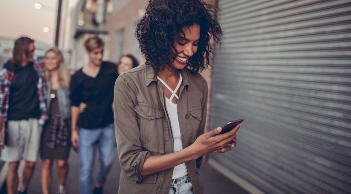 Junge Frau schreibt eine Nachricht am Smartphone, während ihre Freunde hinter ihr stehen.
