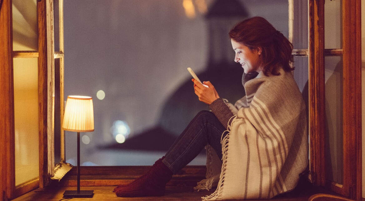 Junge Frau sitzt am Fenster mit ihrem Smartphone und schreibt eine Nachricht.