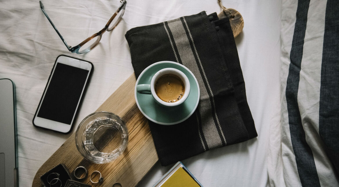Eine volle Espressotasse steht auf dem Bett neben einer Brille und einem Glas Wasser.