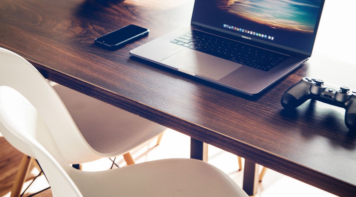 Schwarzer DualShock 4-Controller liegt neben einem Macbook auf dem Schreitisch.
