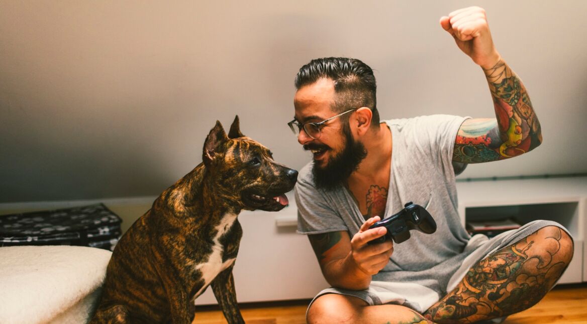 Mann sitzt auf dem Boden und spielt Xbox. Daneben sitzt ein Hund.