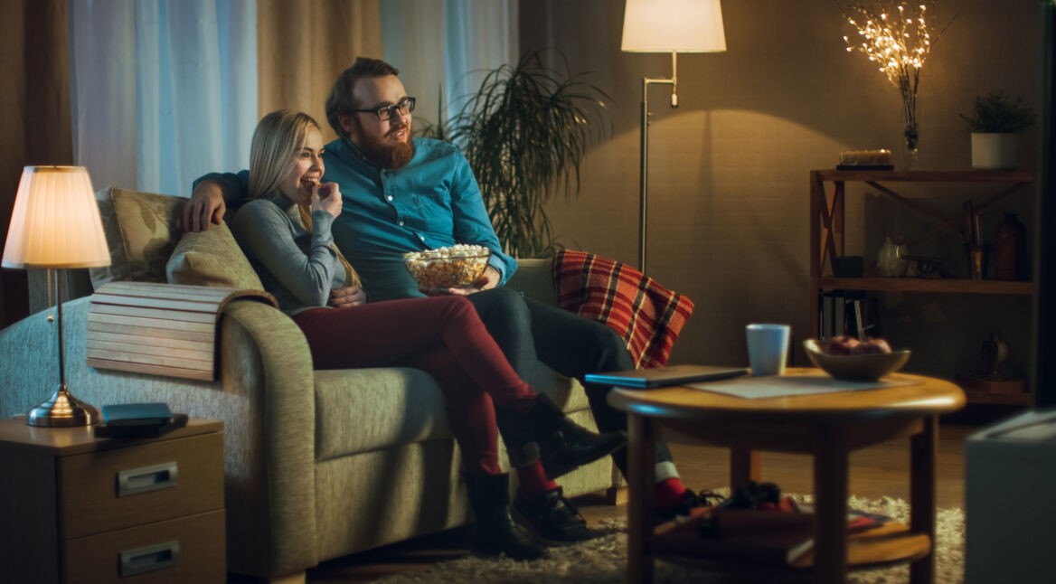 In the Evening Man and Woman are Sitting on the Sofa Watching TV and Eating Popcorn. Living Room is Cozy.