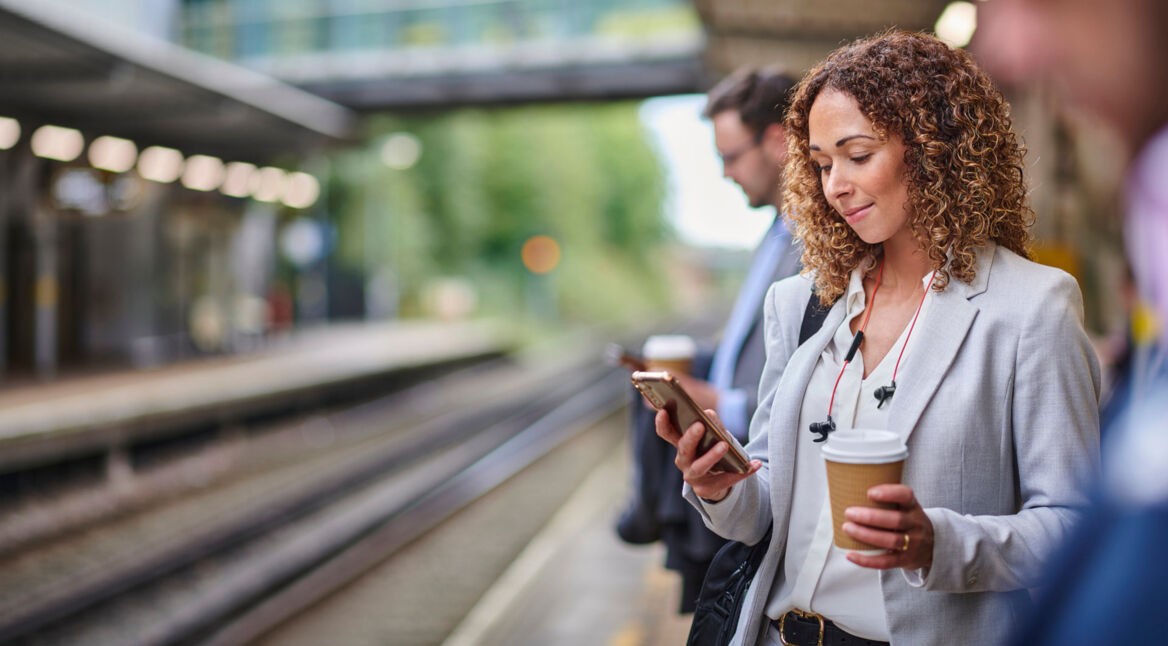 Eine junge Frau steht am Bahngleis und schaut auf ihr Smartphone.