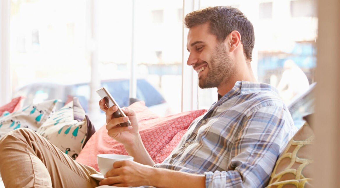 Junger Mann sitzt auf der Couch und schaltet die Akku-Warnung seines Smartphones aus.