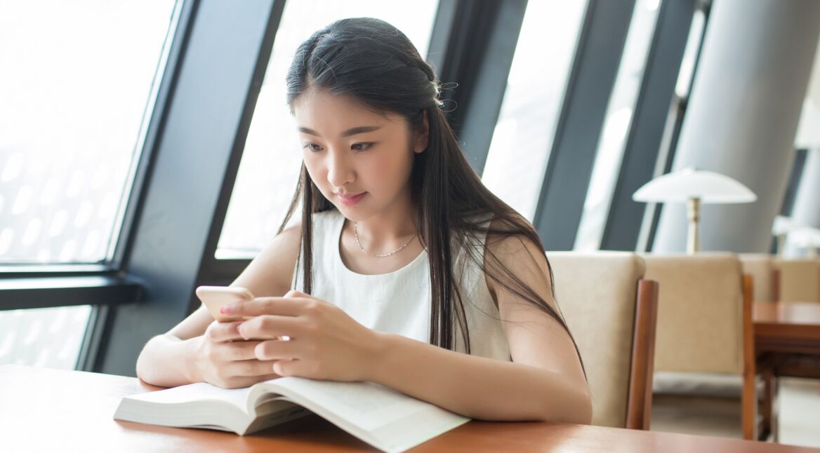 Eine junge Frau lernt mit ihrem Smartphone in der Bibliothek und nutzt dabei den Google Assistant