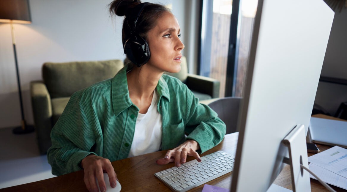 Frau mit Kopfhörern sitzt am Schreibtisch und guckt konzentriert auf den Computer-Bildschirm.