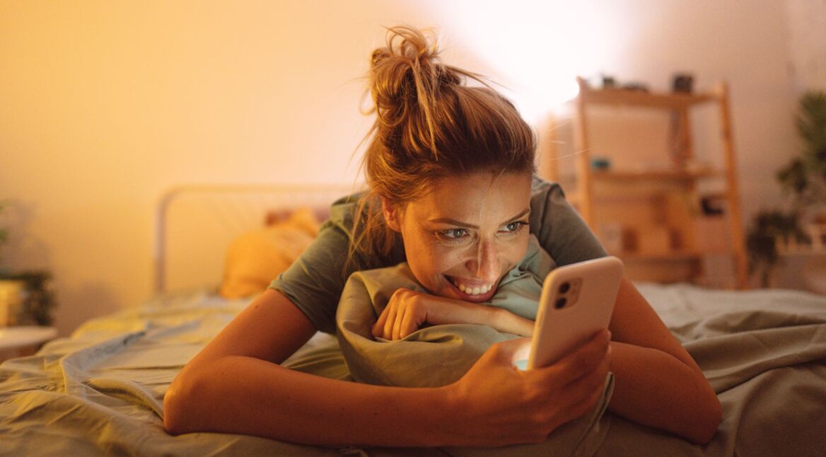 Frau liegt auf dem Bett und schaut aufs Smartphone.