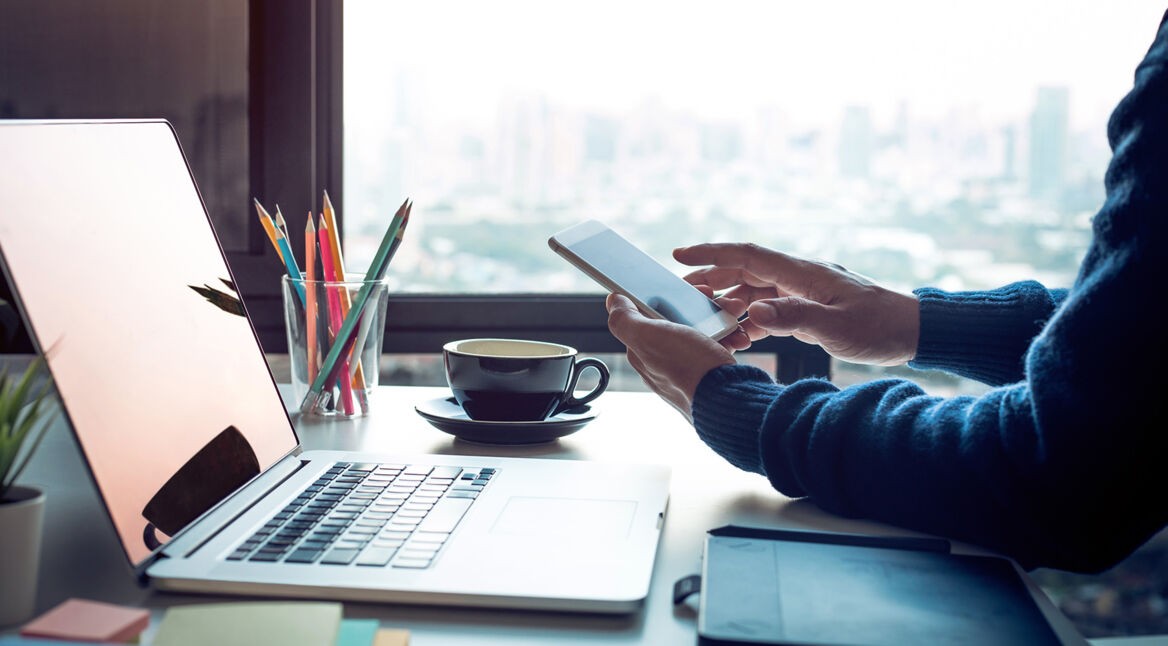 Person mit Handy in der Hand sitzt vor einem Laptop