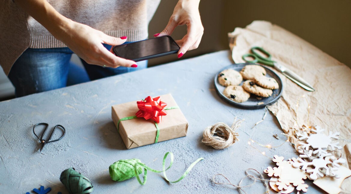 Eine Frau fotografiert ein von ihr verpacktes Geschenk mit dem Smartphone auf einem Basteltisch.