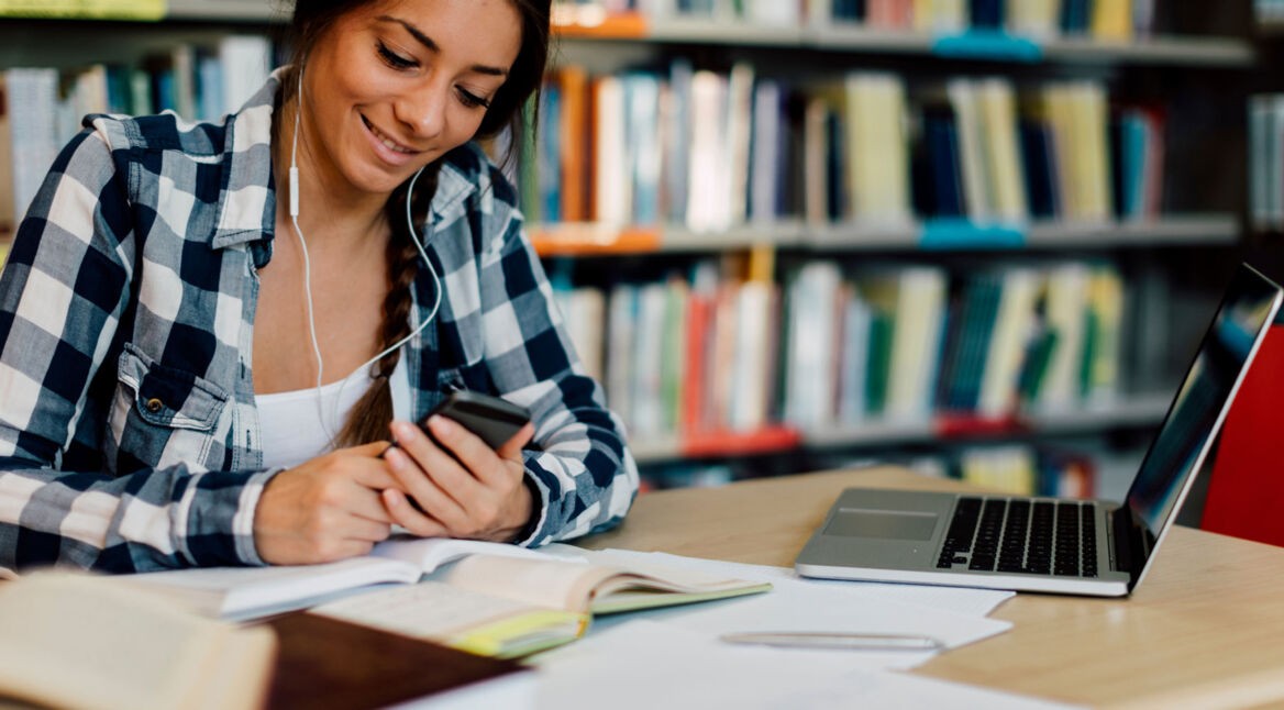 Junge Frau in Bibliothek lernt mit Laptop und am Smartphone