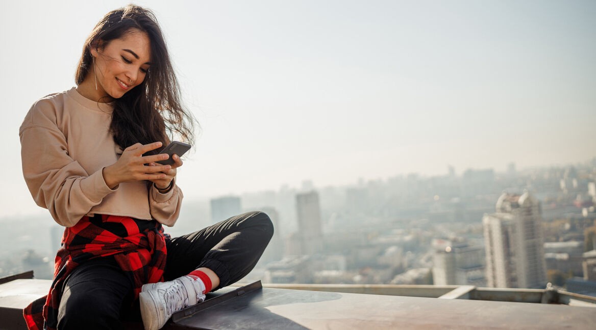 Frau sitzt auf einer Mauer und schaut auf ihr Handy.