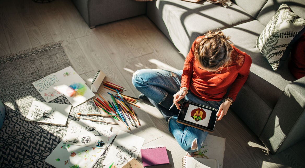 Eine Frau sitzt auf dem Wohnzimmerboden und zeichnet via Tablet.