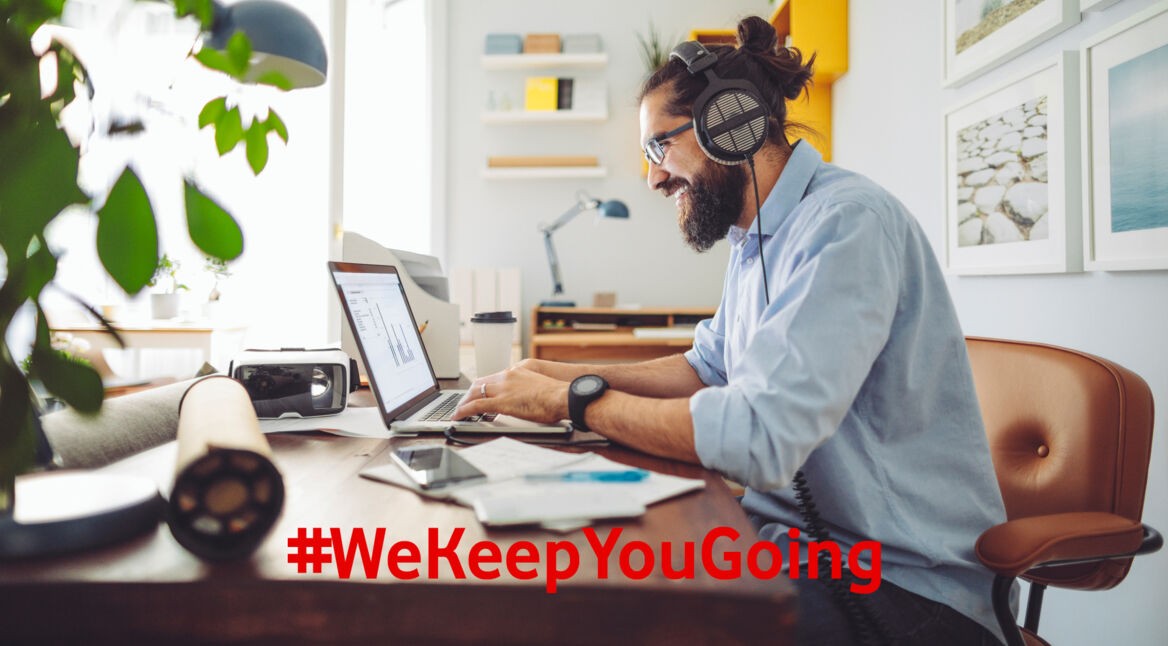 Ein junger Mann trägt Kopfhörer beim Arbeiten am Laptop zuhause und lacht dabei.