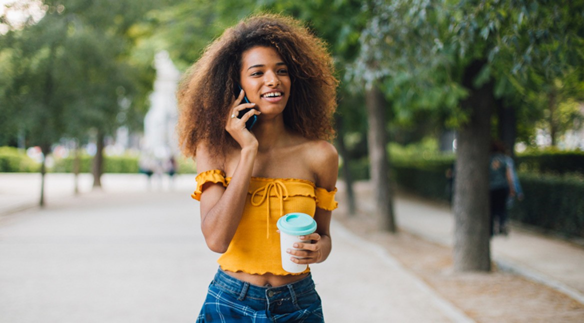 Junge Frau in gelbem Oberteil spaziert durch Park und telefoniert dabei mit Vodafone Crystal Clear