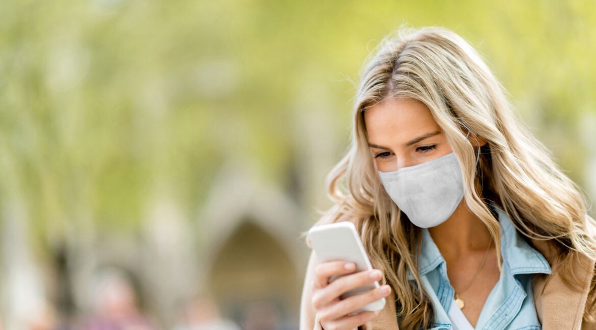 Eine junge Frau trägt einen Mundnasenschutz und schaut auf ihr Smartphone.