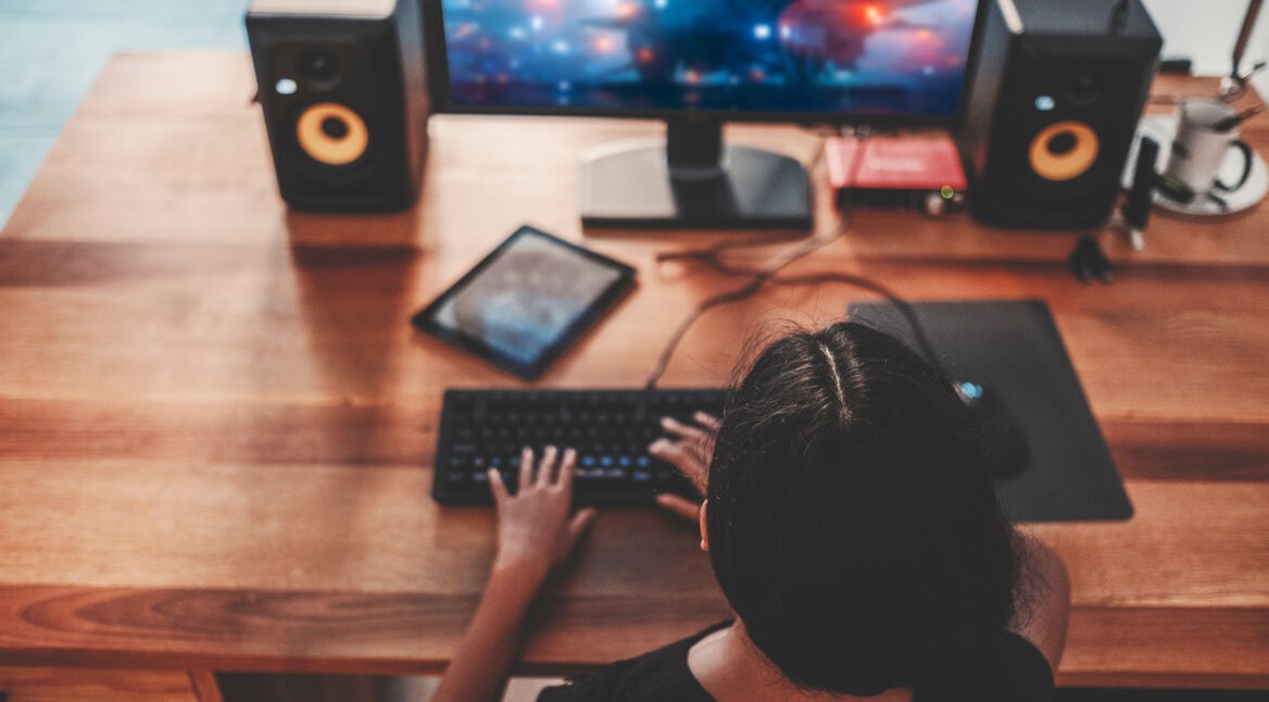 Eine junge Frau spielt ein Spiel am Computer zuhause und bedient dazu ihre Tastatur.