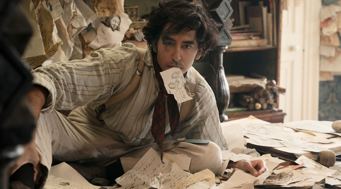 Dev Patel als David Copperfield im gleichnamigen Film inmitten von Papierstapeln, einen trägt er im Mund.