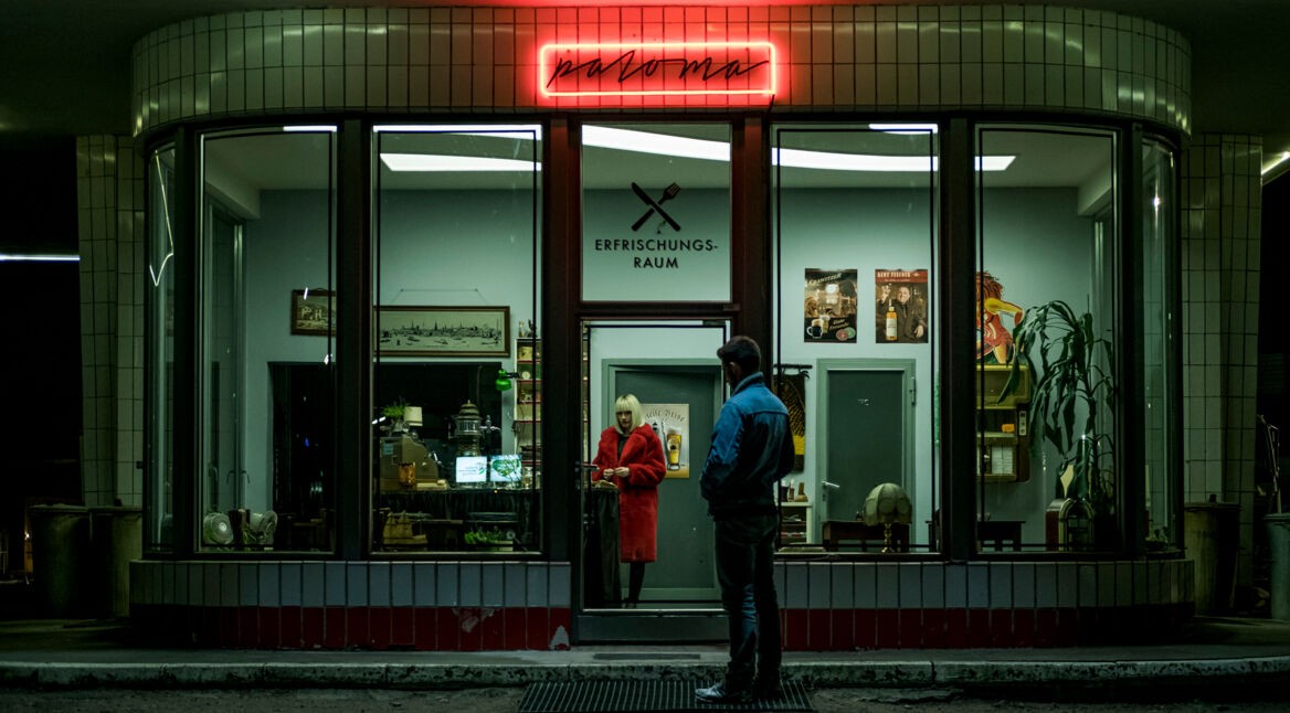 Ein junger Mann und eine Frau mit rotem Mantel stehen vor bzw. in einem Erfrischungsraum mit roter Leuchtschrift.