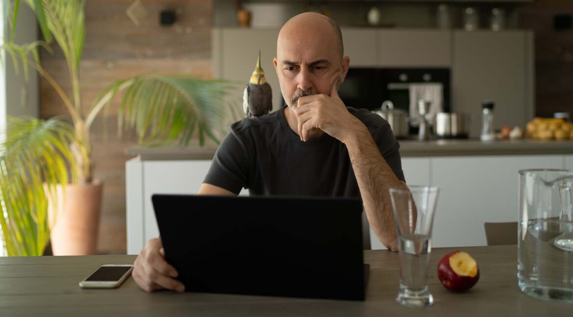 Mann sitzt zu Hause in der Küche am Laptop und grübelt