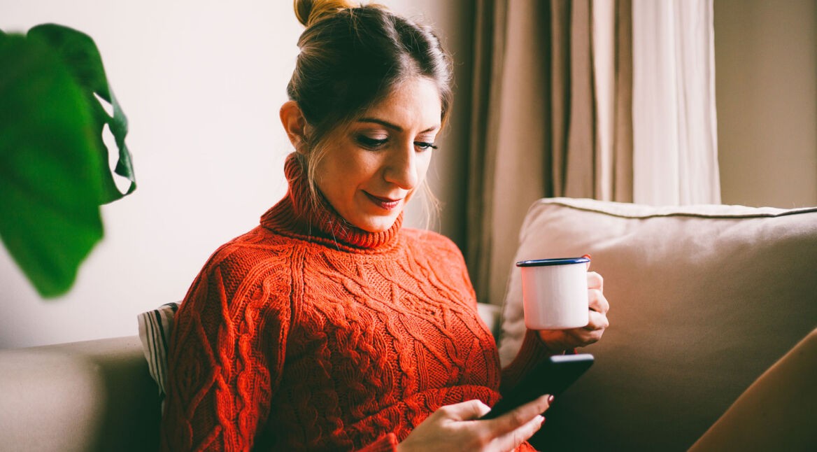 Eine Frau liegt mit einem Heißgetränk auf der Couch und surft auf ihrem Smartphone.