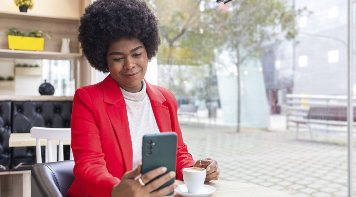 Eine Frau in leuchtend rotem Blazer schaut auf ihr Smartphone, während sie eine Tasse Kaffee in einem Café genießt.