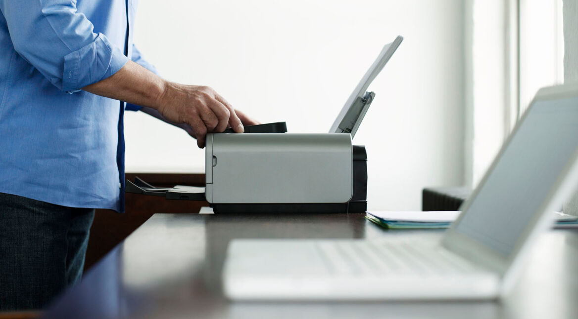 Laptop im Vordergrund, im Hintergrund arbeitet ein Mann am Drucker