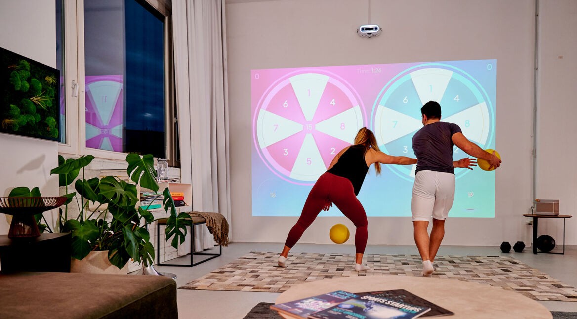 Eine Frau und ein Mann spielen ein Ballspiel mit der Fitness-Konsole Limbic Active gegeneinander, indem sie die Bälle an die Wand schmeißen.