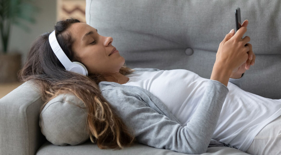 Frau mit Bluetooth-Kopfhörern liegt auf dem Sofa und nimmt Einstellungen am Handy vor