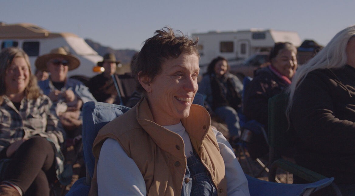 Fern (Frances Dormand) sitzt lachend mit Nomad:innen draußen auf einem Klappstuhl.