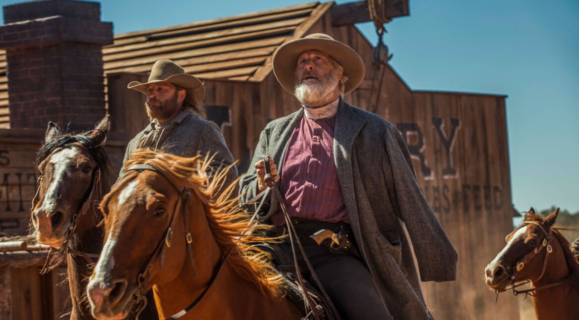 Im Artikel zu den besten Western-Serien siehst du ein Bild aus der Serie Godless ist Jeff Daniels als Frank Griffin. Er reitet auf einem braunen Pferd durch eine staubige Westernstadt, begleitet von einem weiteren Reiter