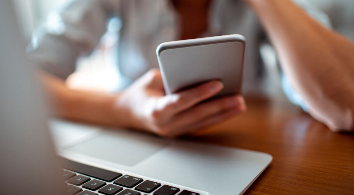 Frau hält Smartphone in der Hand, während sie am Laptop sitzt